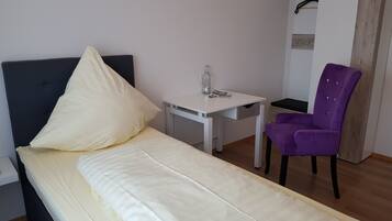 A single bed with a dark headboard, a small white desk with a bottle and glasses, a purple chair, and a wardrobe with a wooden accent.