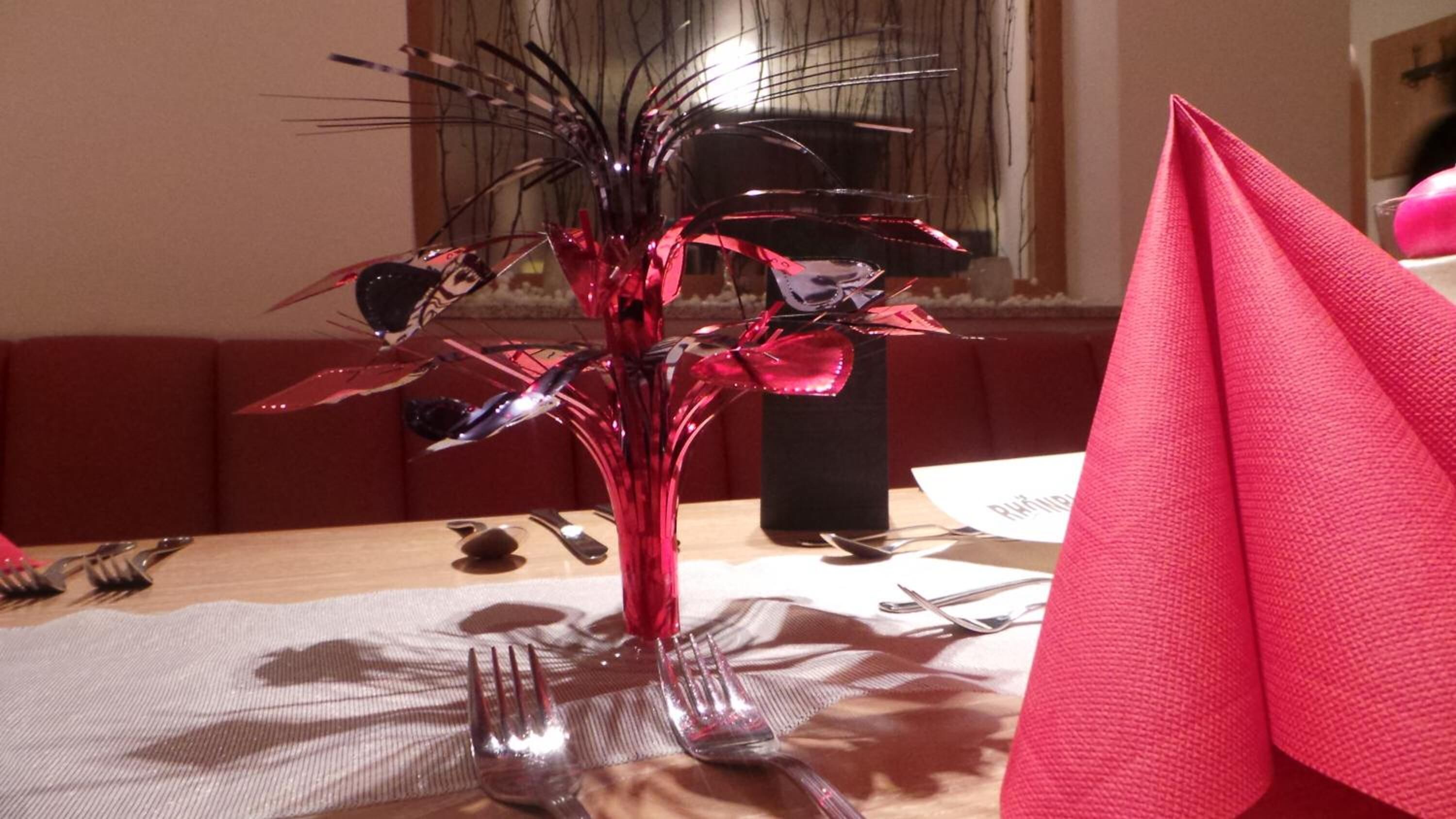 Una mesa de comedor con un centro de mesa en un florero rojo, una servilleta rosa y cubiertos.