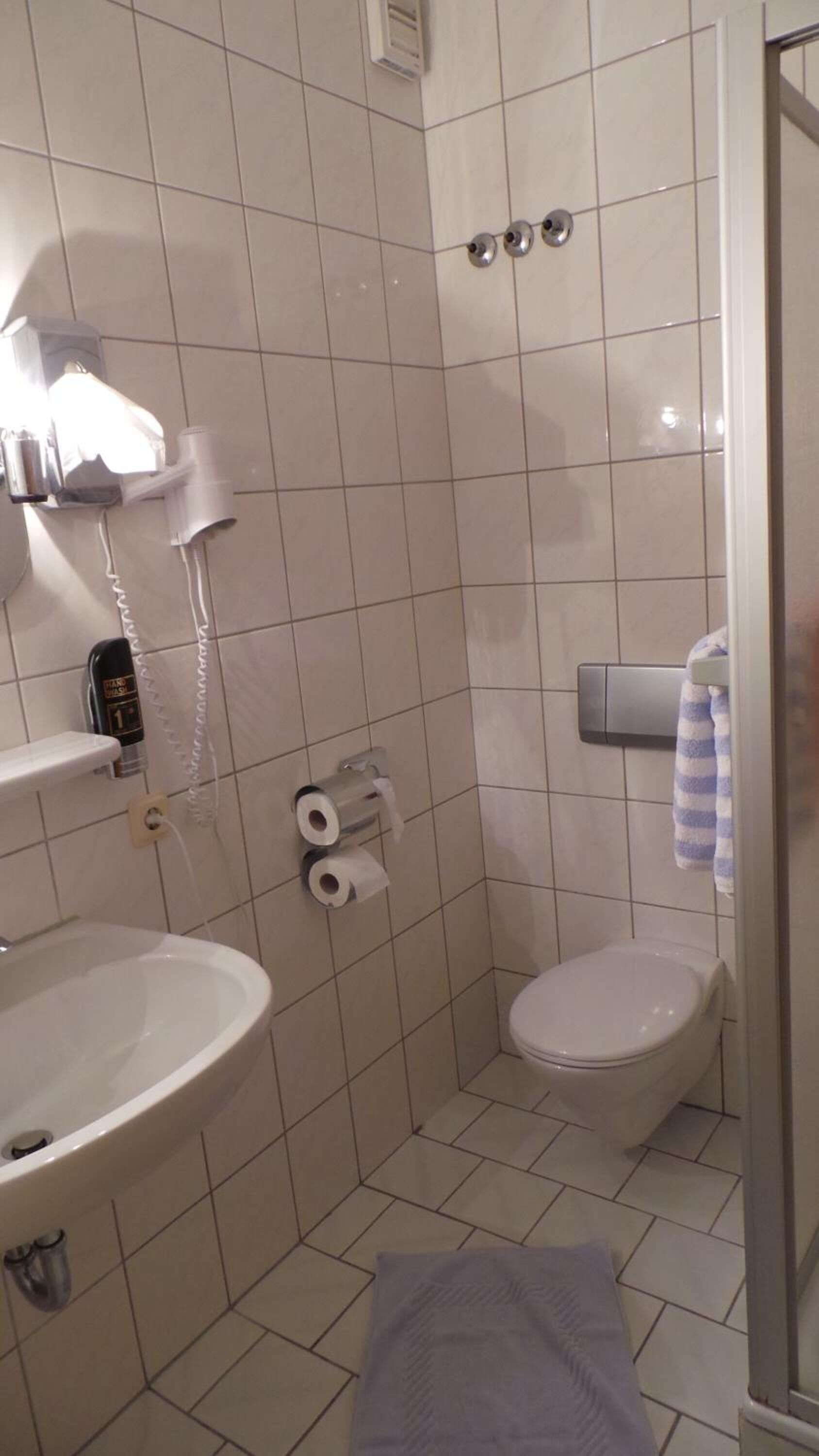 Un baño pequeño y limpio con lavamanos, inodoro y ducha. Hay dos rollos de papel higiénico y una toalla colgada en la pared.