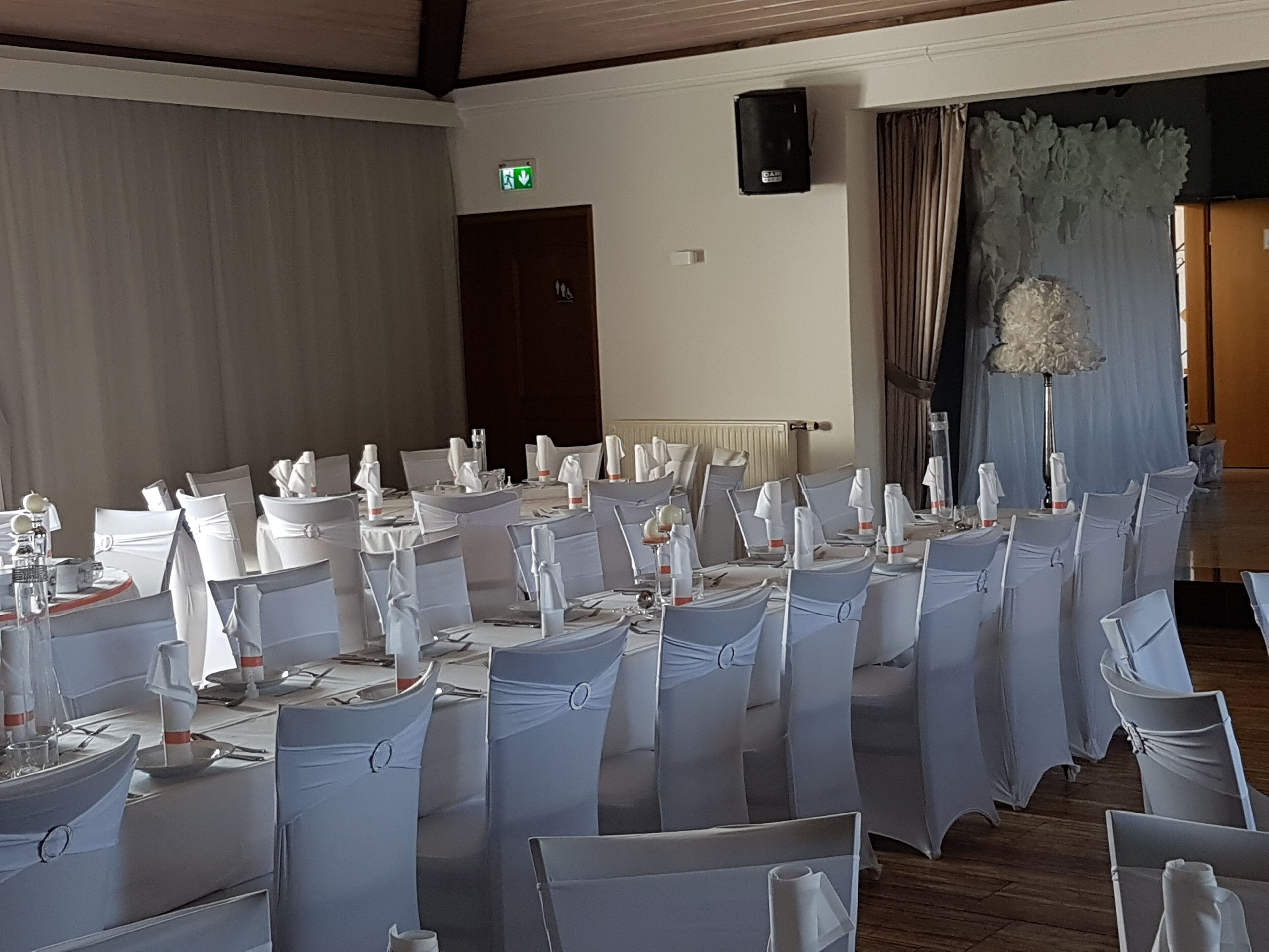 Un salón de banquetes con mesas redondas dispuestas para un evento formal, sillas blancas con cintas, un escenario con cortinas y un altavoz montado en la pared.
