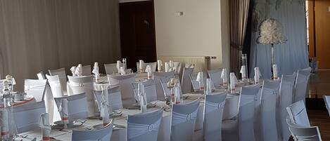 A banquet hall with round tables set for a formal event, white chairs with sashes, a stage with curtains, and a speaker mounted on the wall.