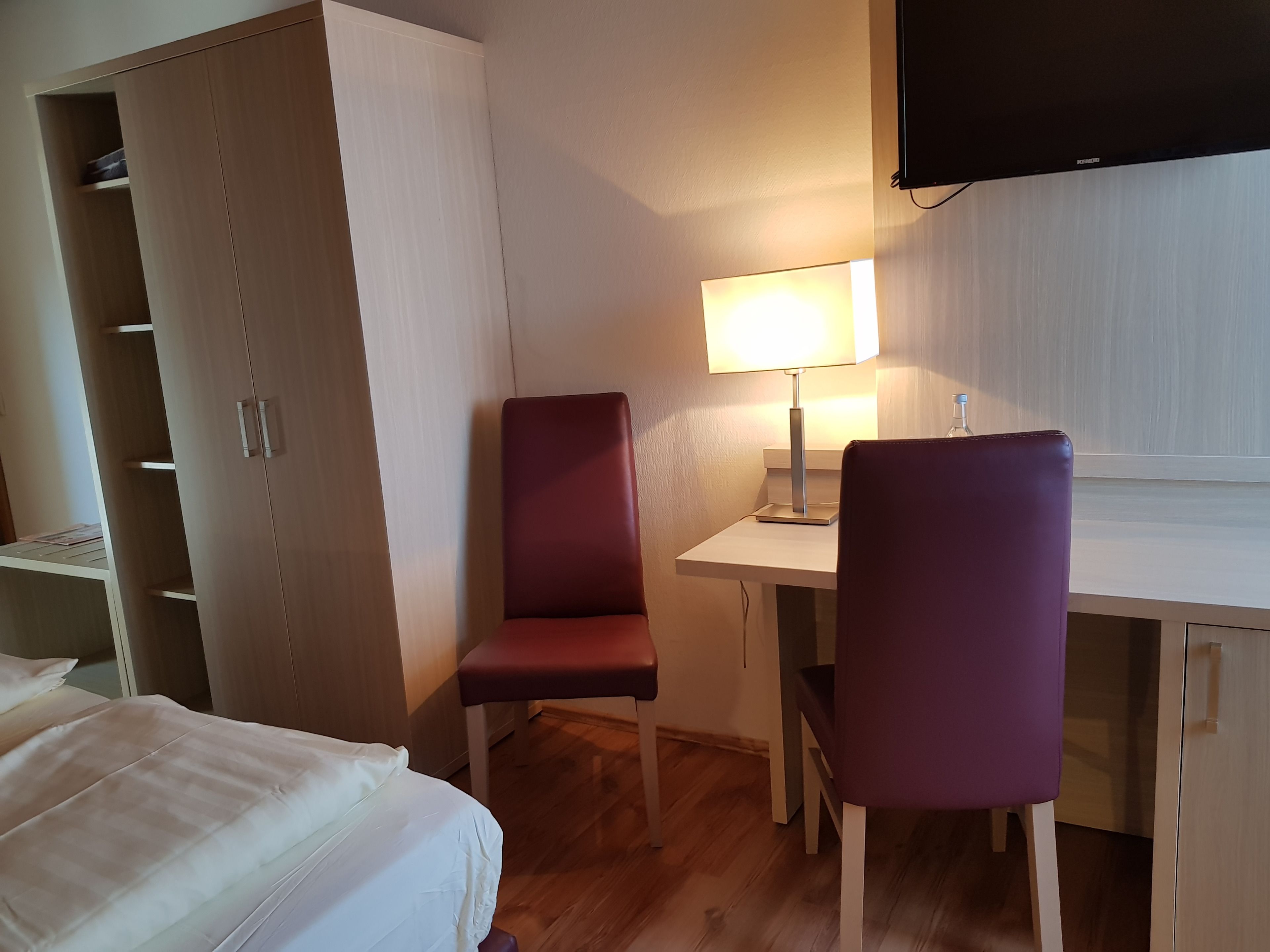 A hotel room with a bed, a desk with a lamp, two chairs, and a wardrobe.