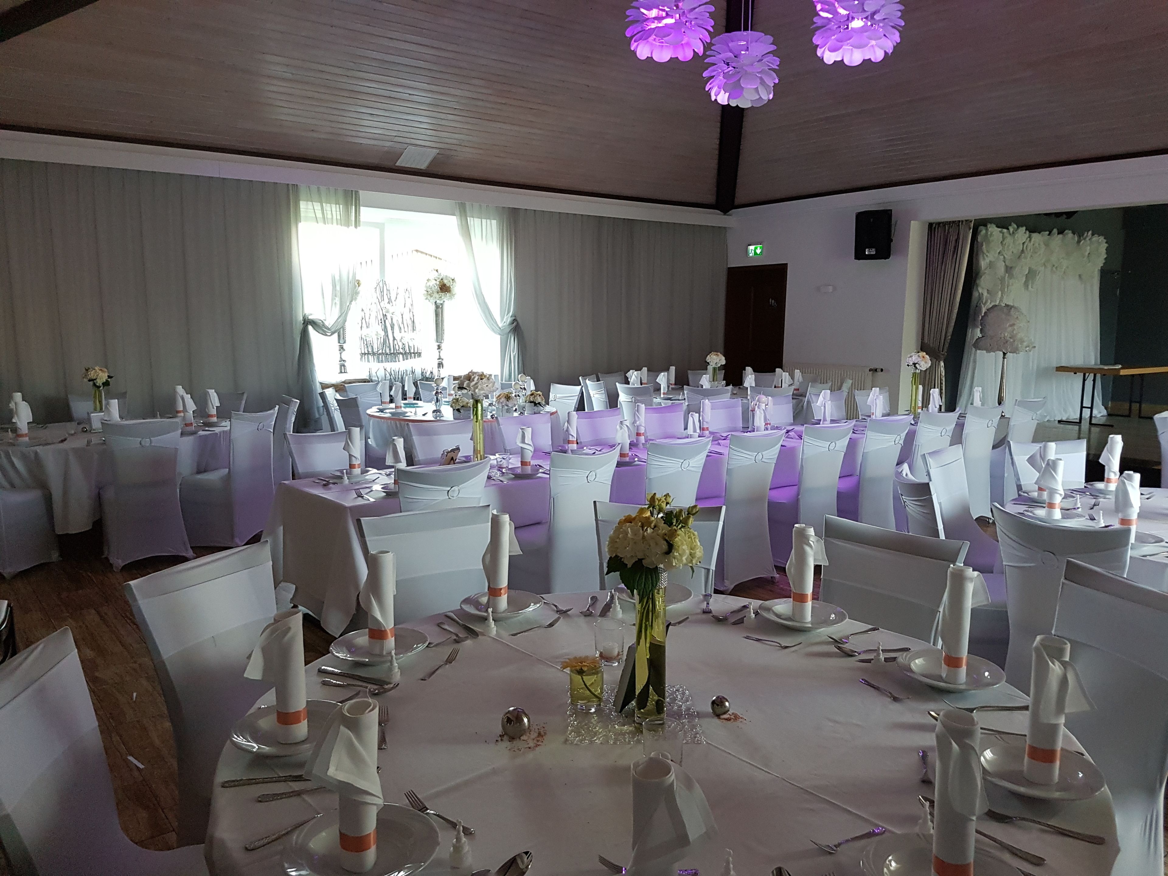 Ein Festsaal mit runden Tischen, gedeckt für eine formelle Veranstaltung, weiße Stühle und violette Beleuchtung.