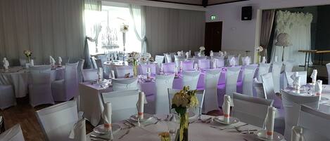 Un salón de banquetes con mesas redondas dispuestas para un evento formal, sillas blancas e iluminación morada.