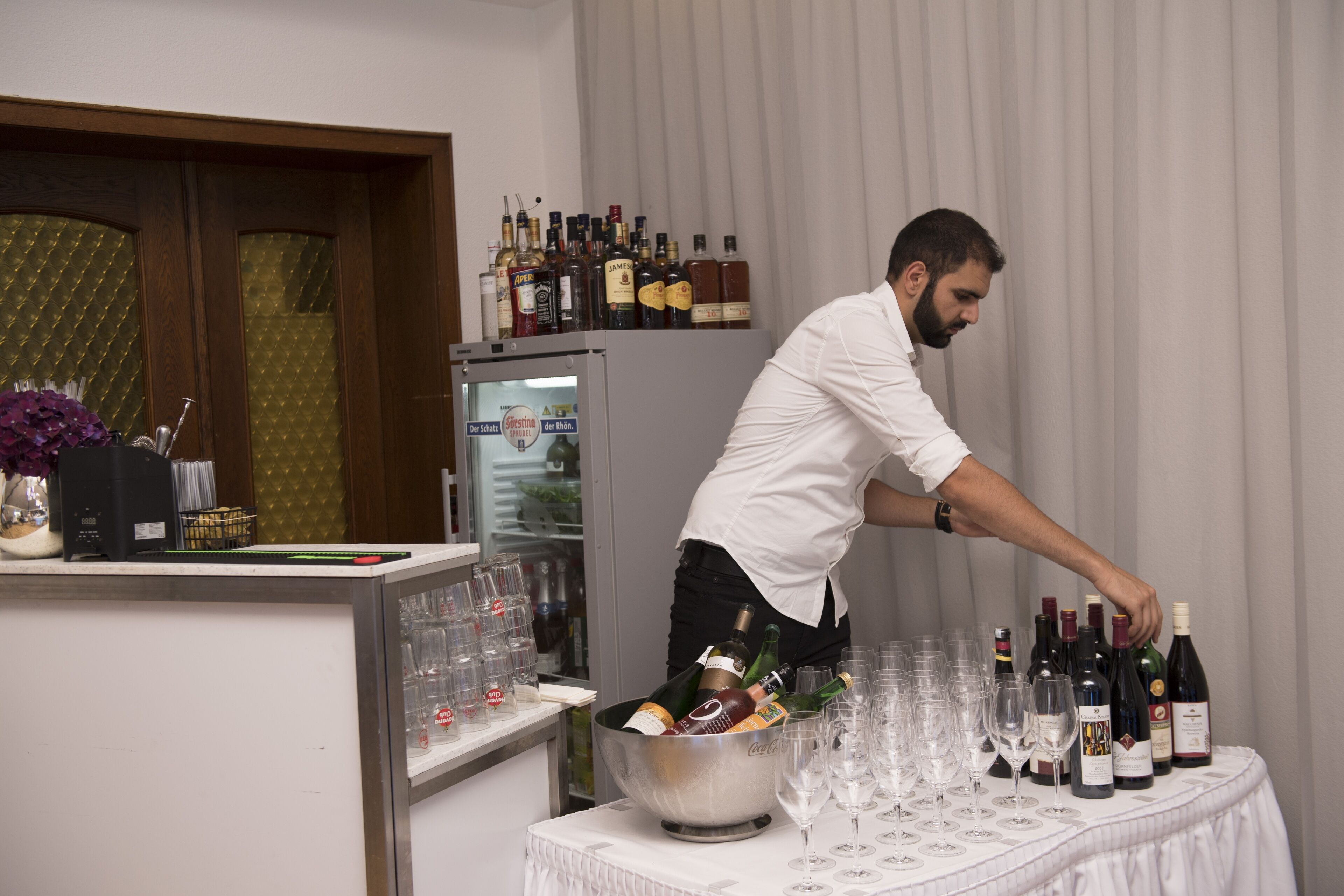 Un espace bar avec un barman préparant des boissons, un réfrigérateur rempli de bouteilles et une table avec des verres et des bouteilles.
