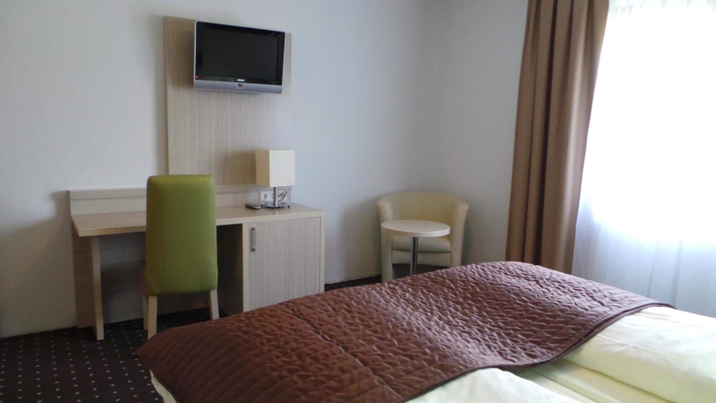 Habitación de hotel con cama, escritorio, silla, televisor y ventana con cortinas.