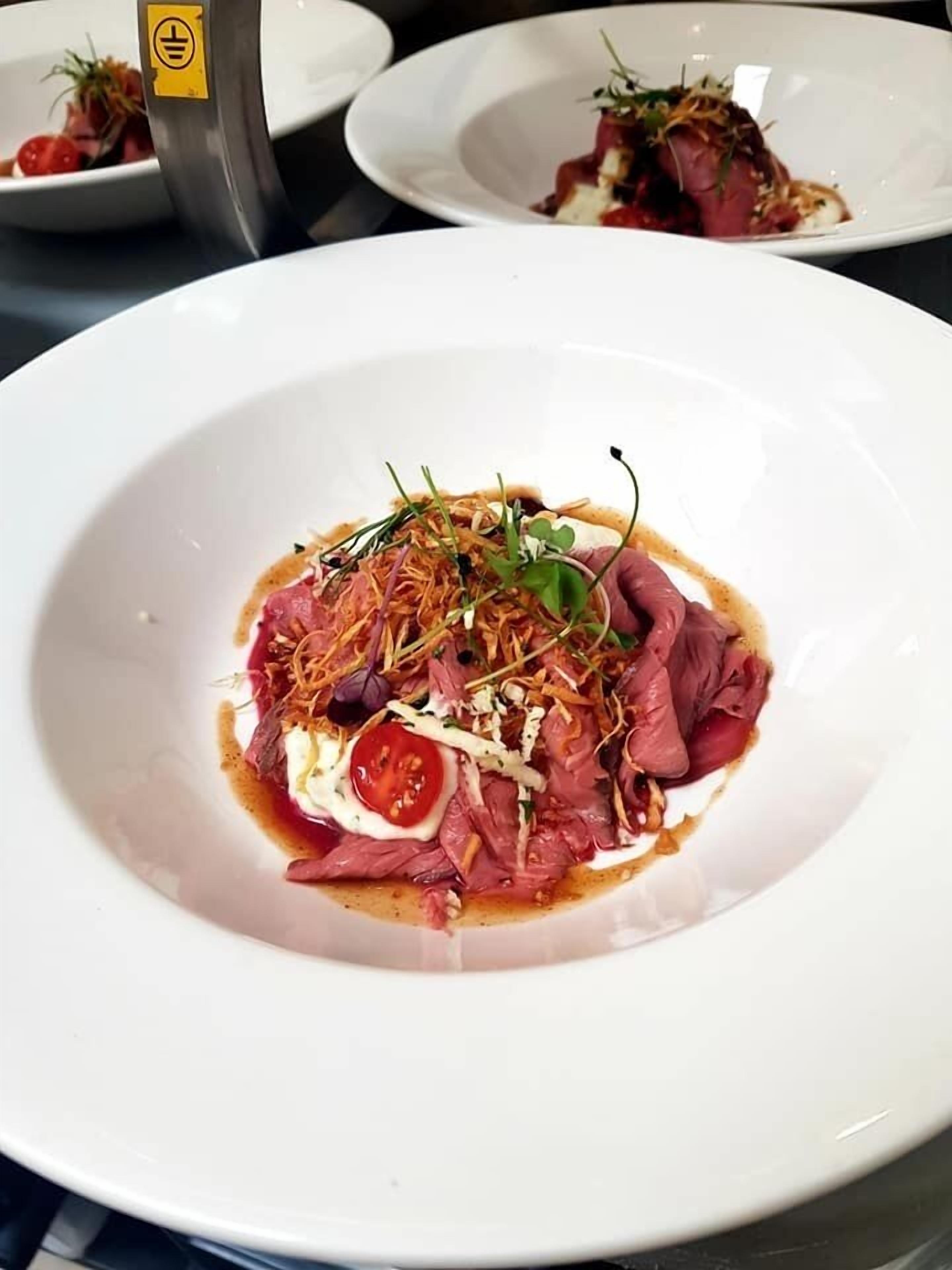 Un plato blanco con guarnición de carne cruda, hierbas y una rodaja de tomate.