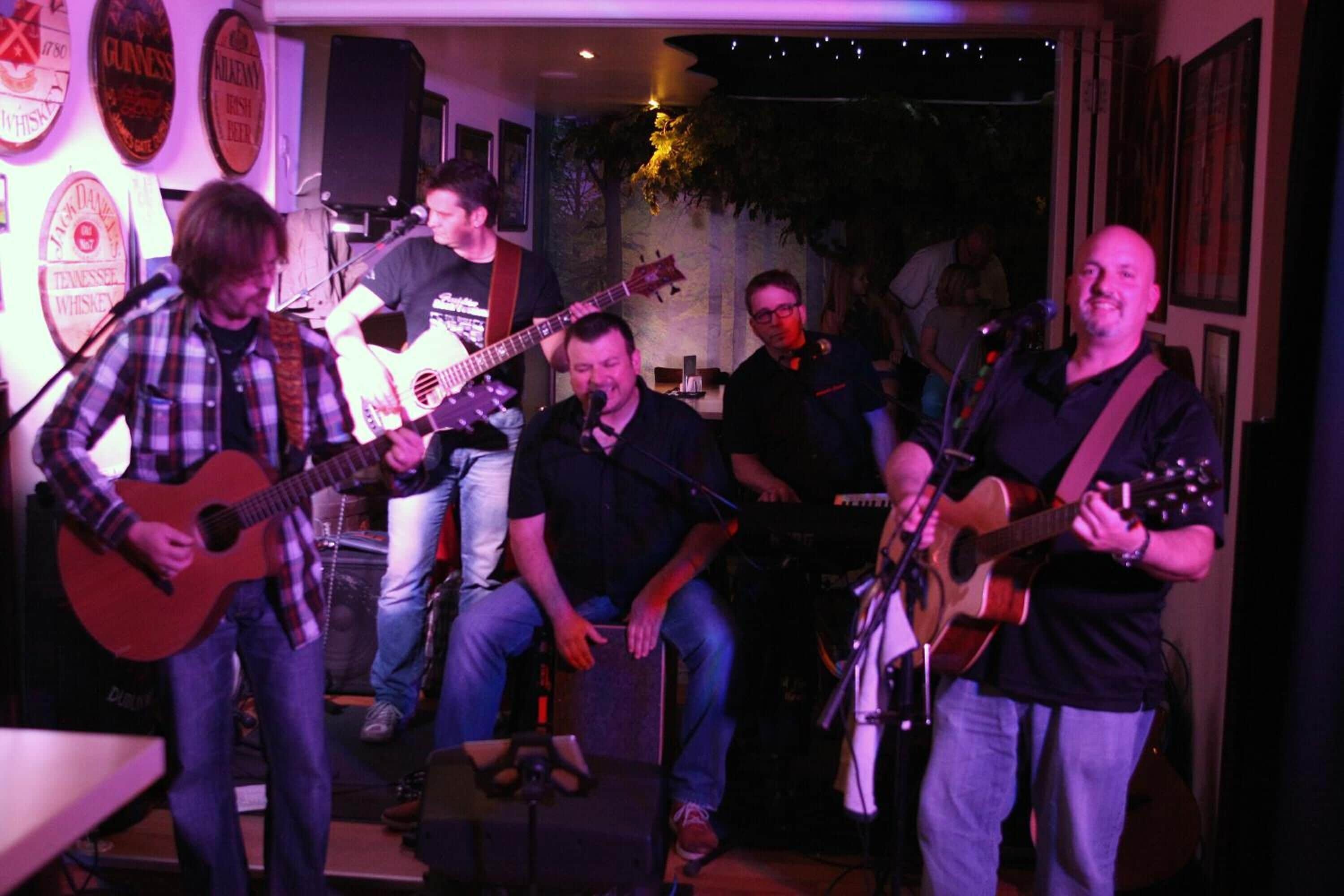 A group of musicians performing in a room with a stage, a microphone stand, and a guitar.