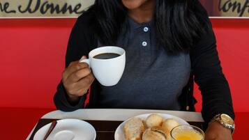Una persona sentada a una mesa con una comida que incluye pan, fruta, un tazón de cereal, un vaso de jugo de naranja y una taza de café.
