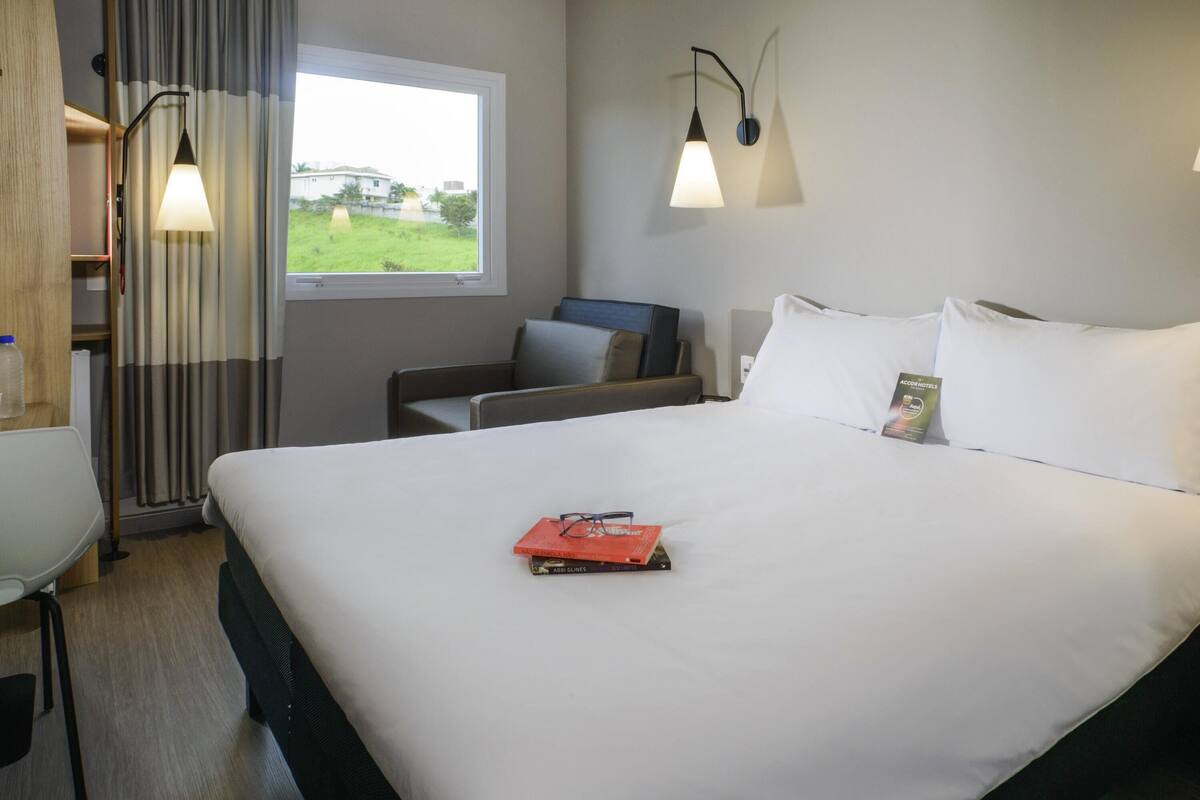 A hotel room with a large bed, a desk with a chair, a window with curtains, and two wall-mounted lamps.