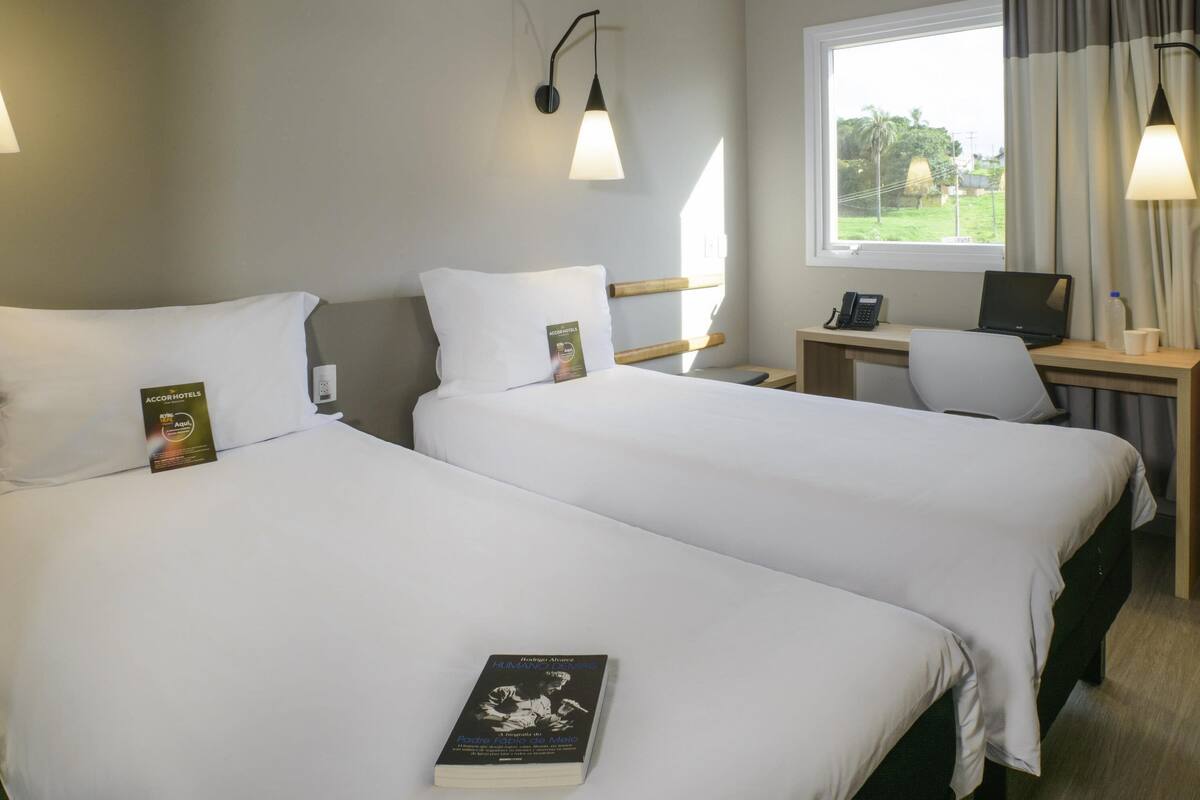 A hotel room with two beds, a desk with a laptop, a phone, and a window with curtains.