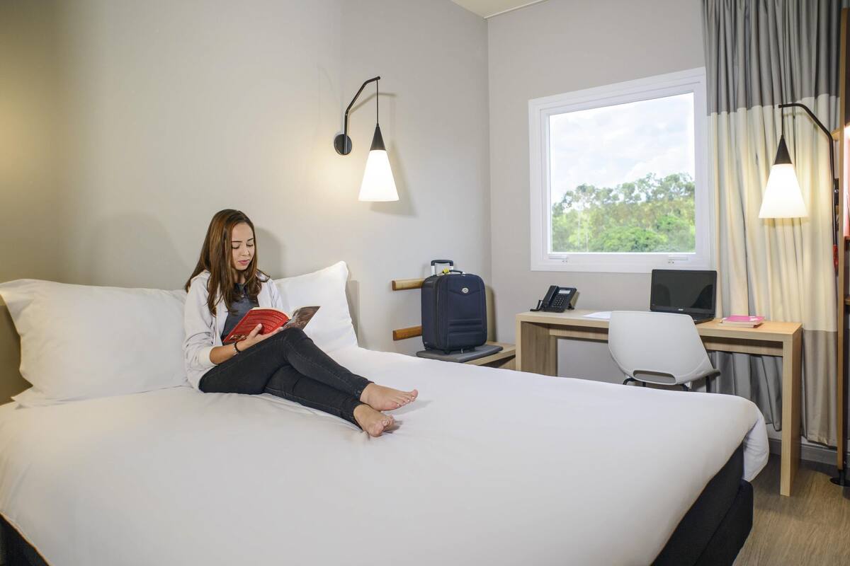 A hotel room with a bed, a desk with a laptop, a suitcase, and a window with a view of greenery.