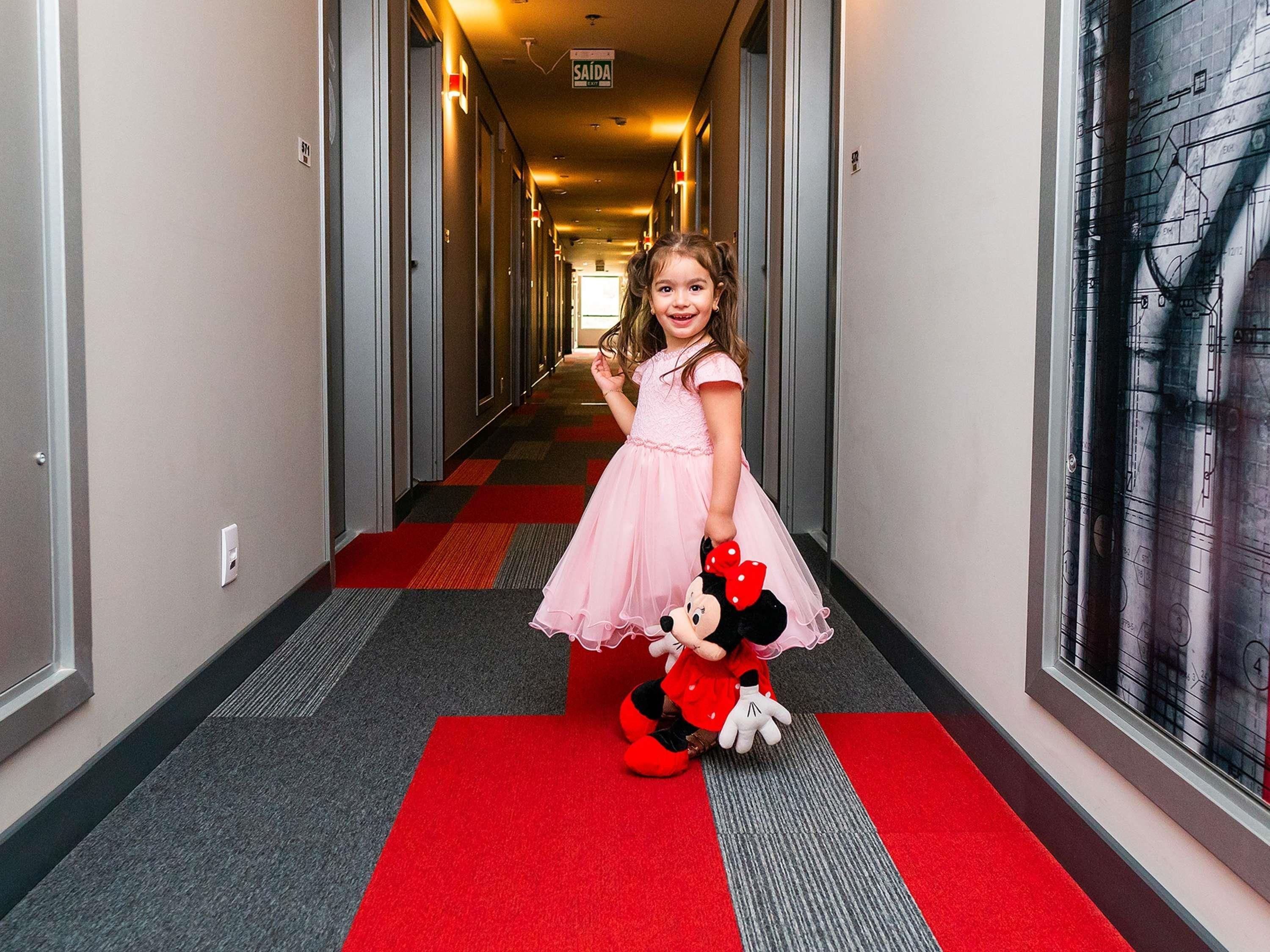 Um corredor de hotel com carpete vermelho, portas em ambos os lados e um brinquedo de pelúcia do Mickey na parede.