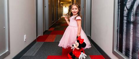 Un couloir d’hôtel avec un tapis rouge, des portes de chaque côté et un jouet en peluche à l’effigie de Mickey Mouse posé sur le sol.
