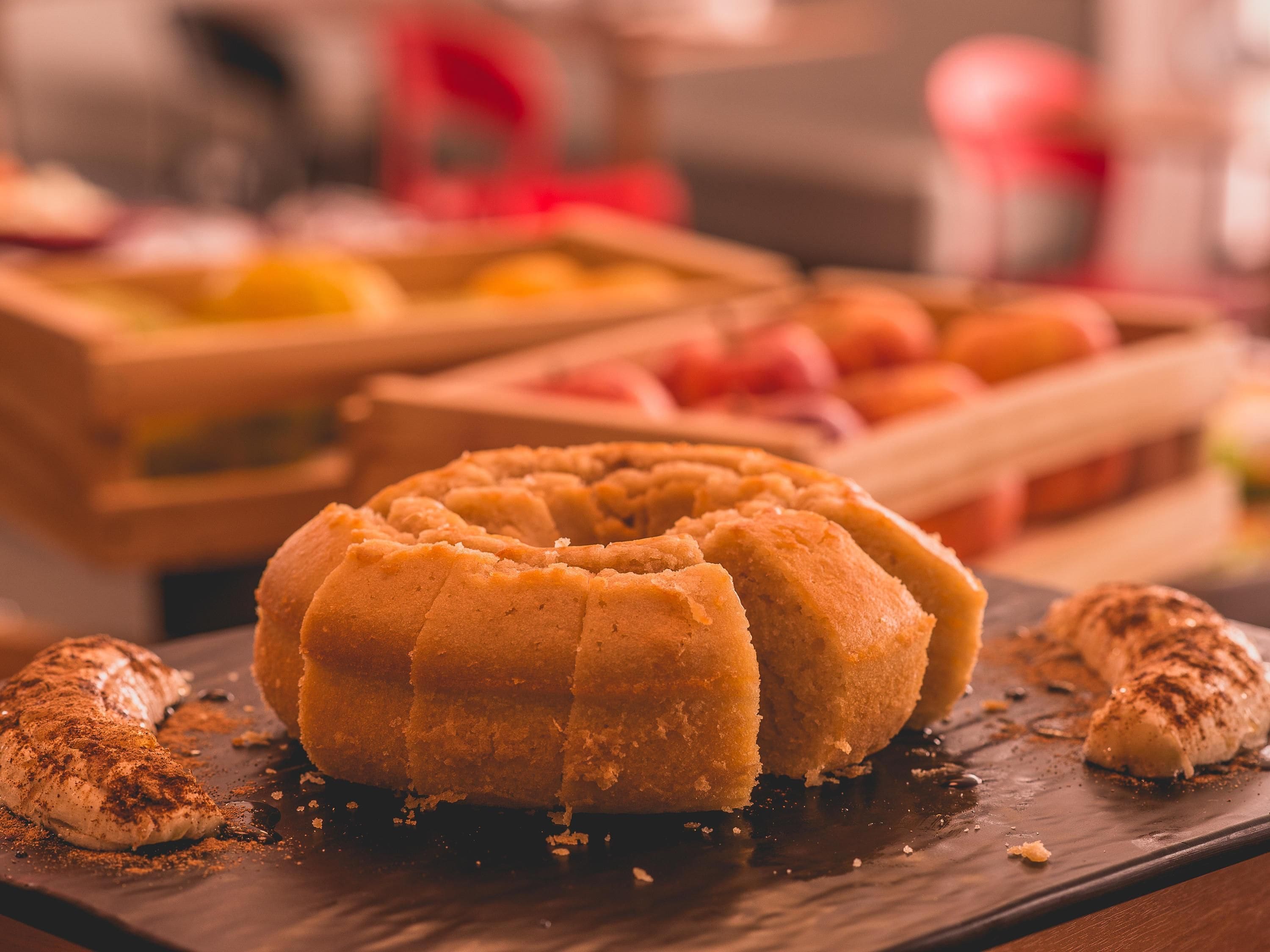 A bundt cake on a dark slate plate with a chocolate drizzle, surrounded by fruits and a blurred background.