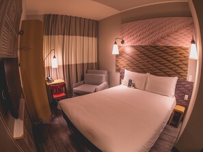 Ein Hotelzimmer mit Bett, Schreibtisch mit Computer, Stuhl und Nachttisch mit Lampe.