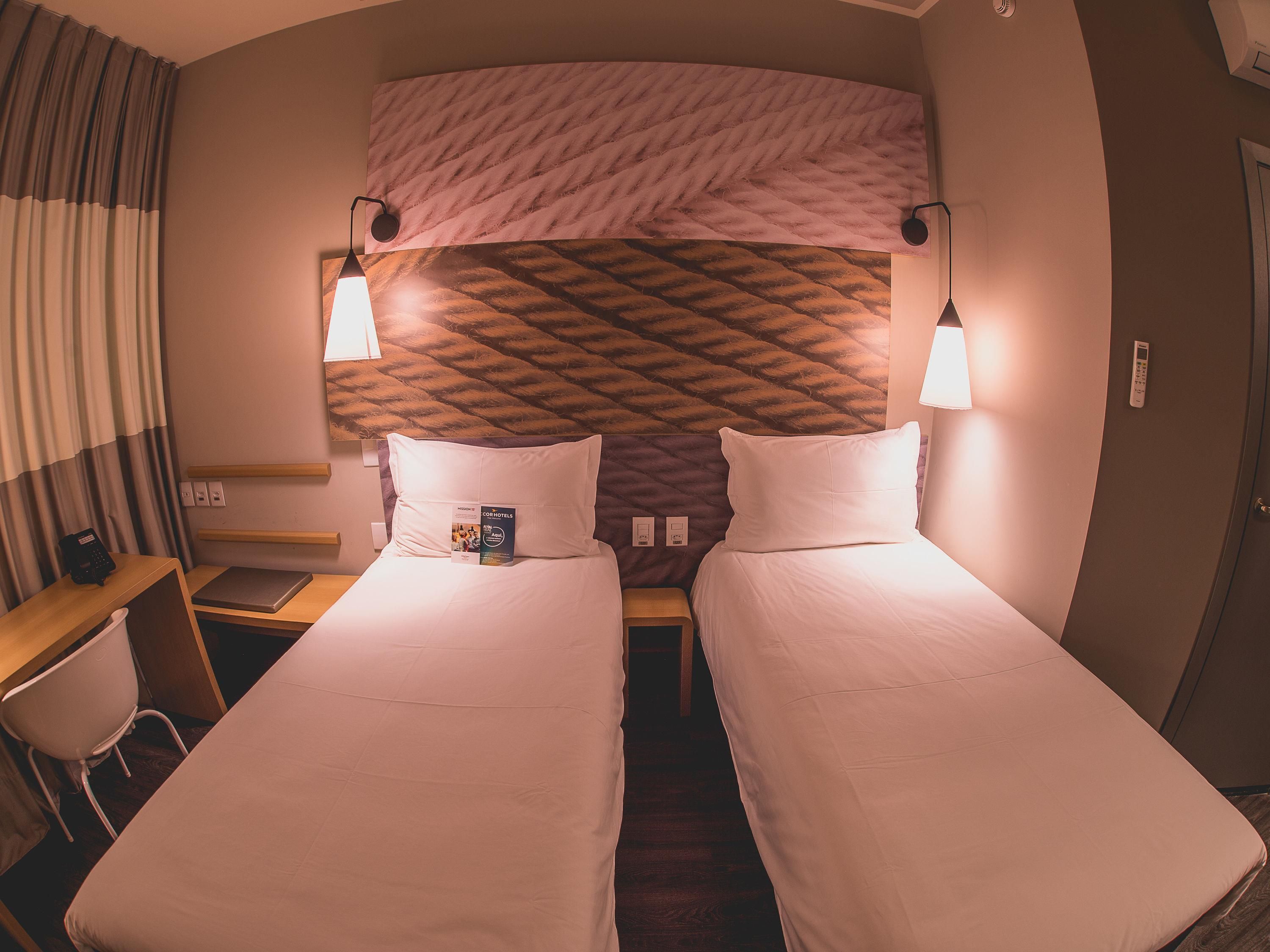 A hotel room with two beds, a desk, a chair, a telephone, and a wall-mounted control panel.