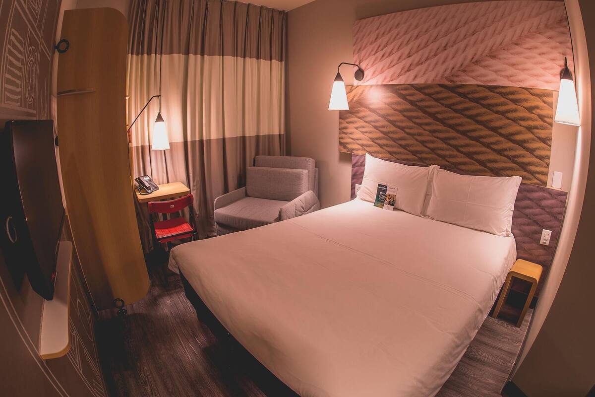 A hotel room with a bed, a desk, a chair, a lamp, and a curtain.
