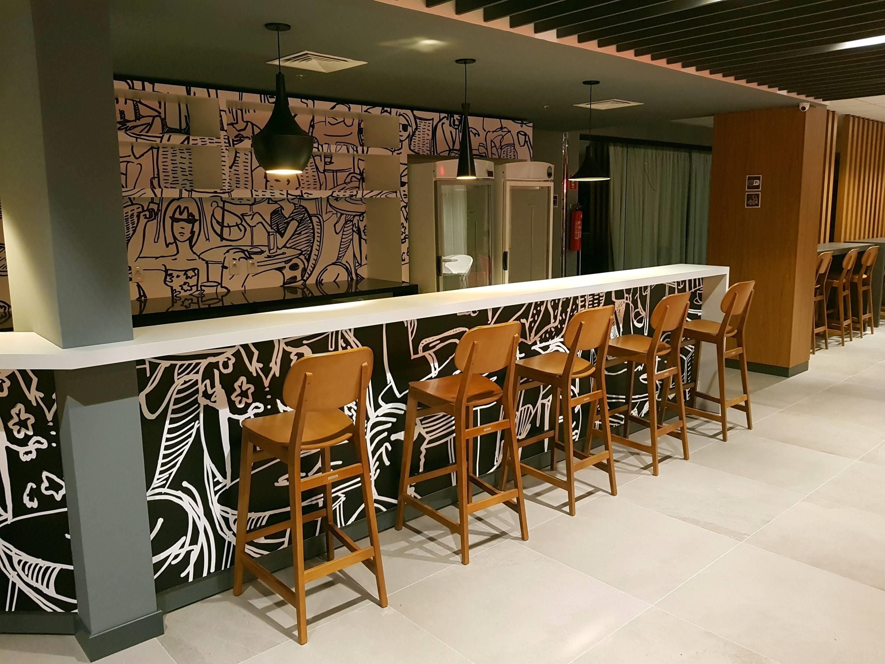 Uma área de bar com um balcão comprido, vários banquinhos de madeira e uma parede com padrões abstratos em preto e branco.