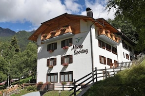 A two-story Alpine Sporting building with wooden balconies and flower boxes, surrounded by lush greenery and mountains.