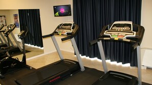 A gym with treadmills and stationary bikes, a TV on the wall, and a dark curtain backdrop. - Hotel Fiore (Fiorenzuola d'Arda)