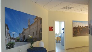 A hotel lobby with a large cityscape painting on the wall, a reception desk, and a dining area with a table and chairs. - Hotel Fiore (Fiorenzuola d'Arda)