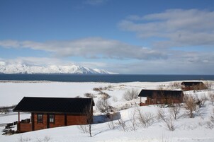 Una cabaña rústica con techo inclinado, rodeada de campos nevados y montañas al fondo.