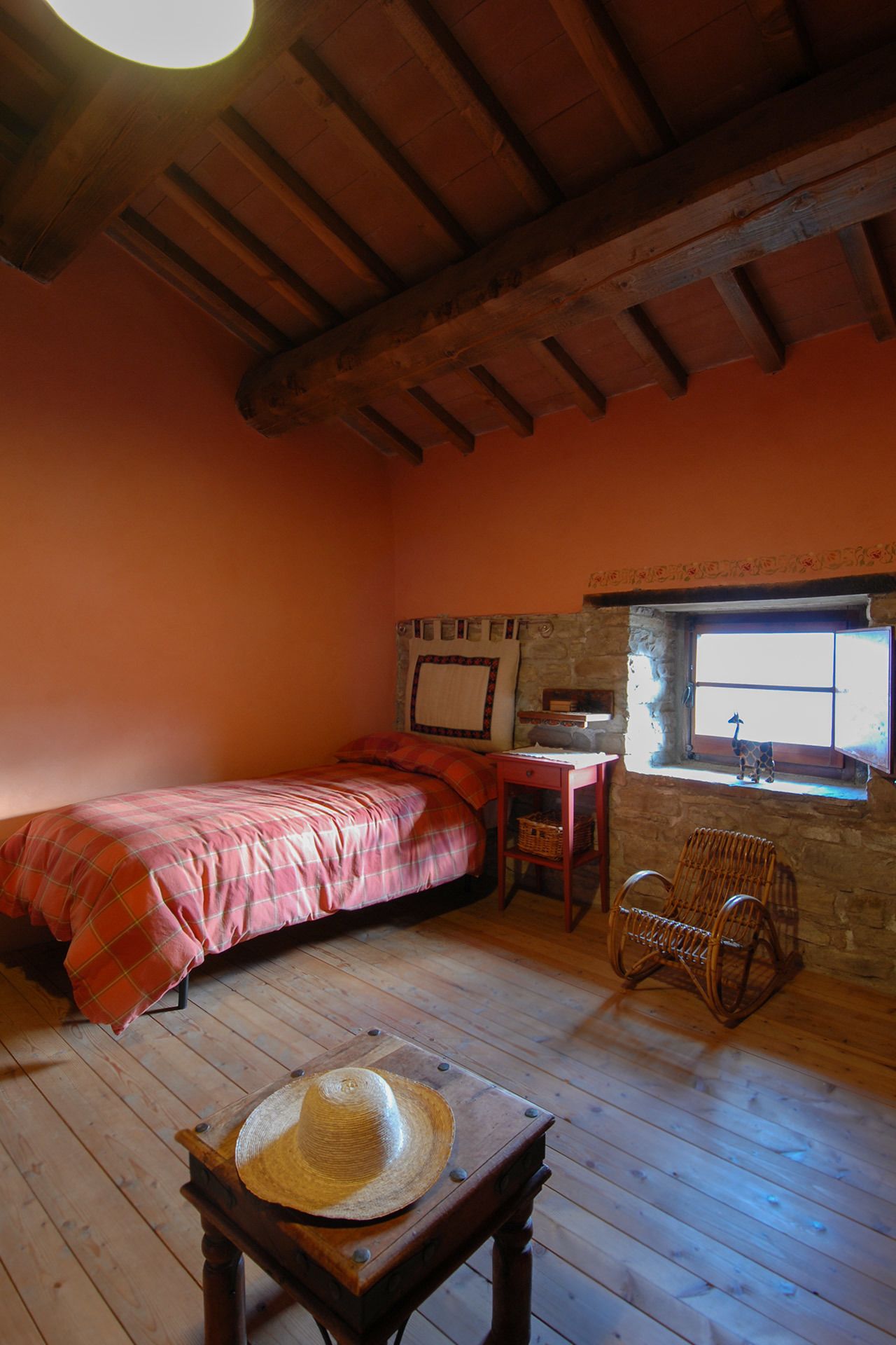 A cozy room with a wooden ceiling, a bed with a plaid blanket, a wicker chair, and a small window with a view.