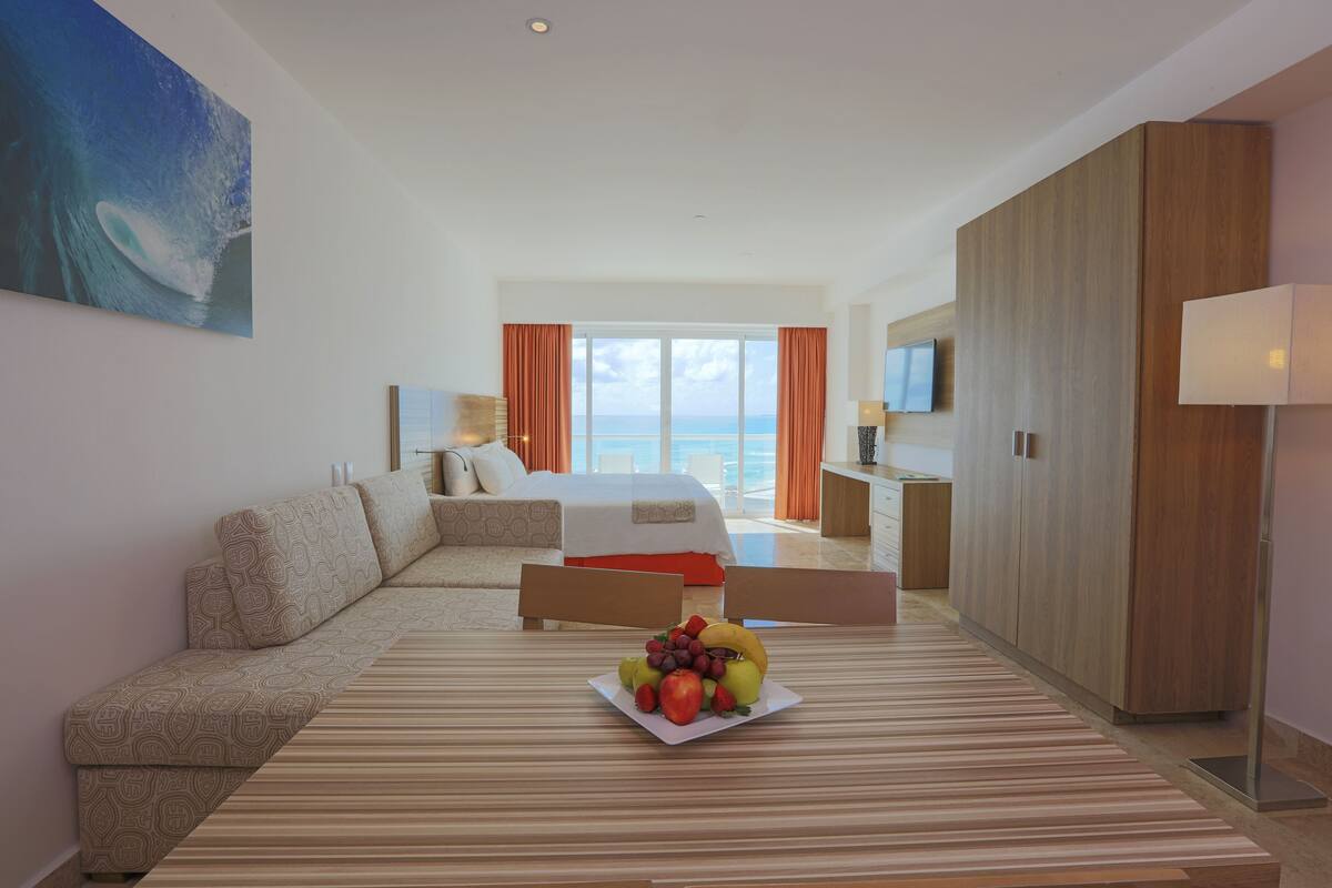 Quarto de hotel com cama, sofá, mesa de jantar com frutas e vista para o mar.