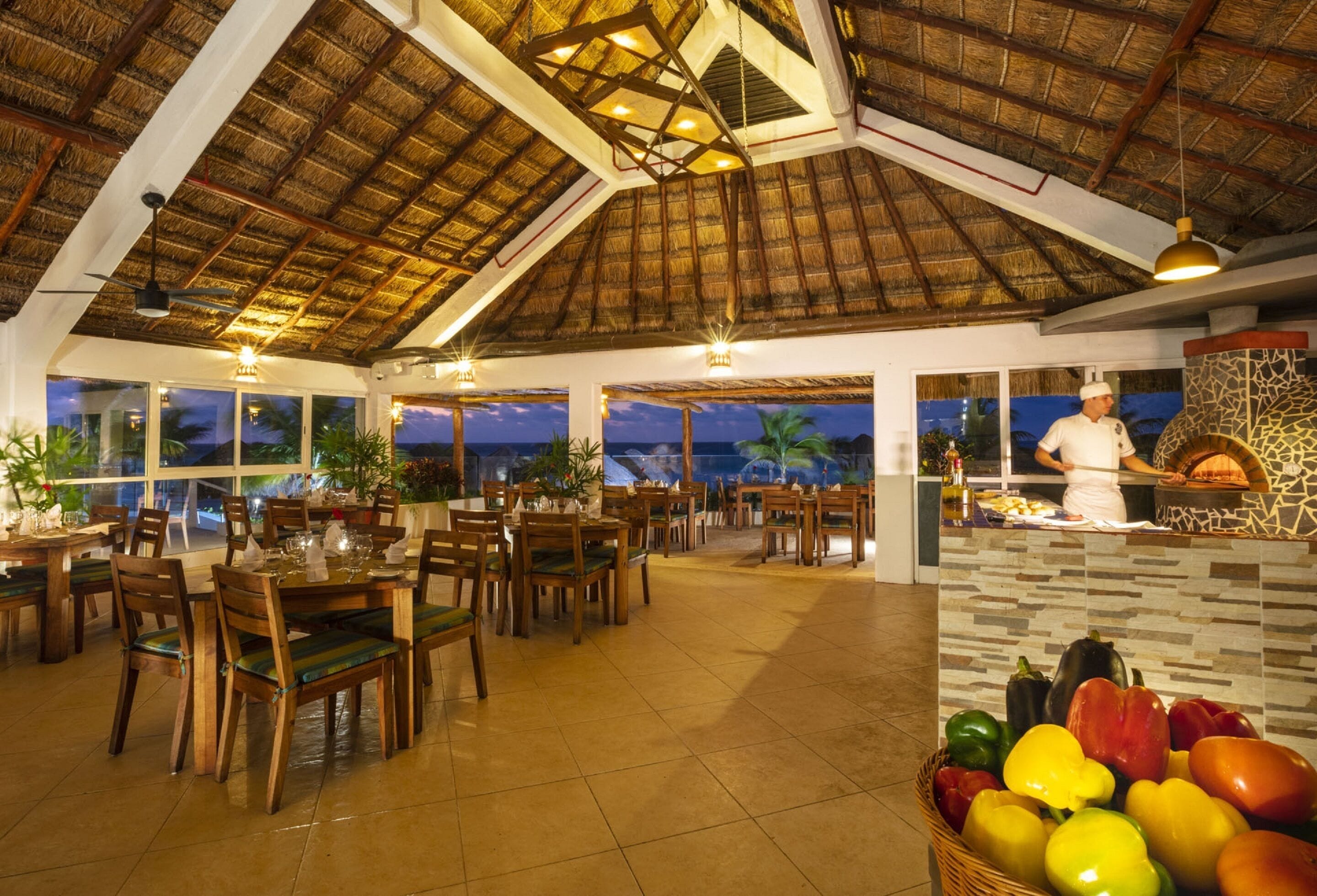 A restaurant with a thatched roof, wooden tables and chairs, and a view of the ocean at dusk.
