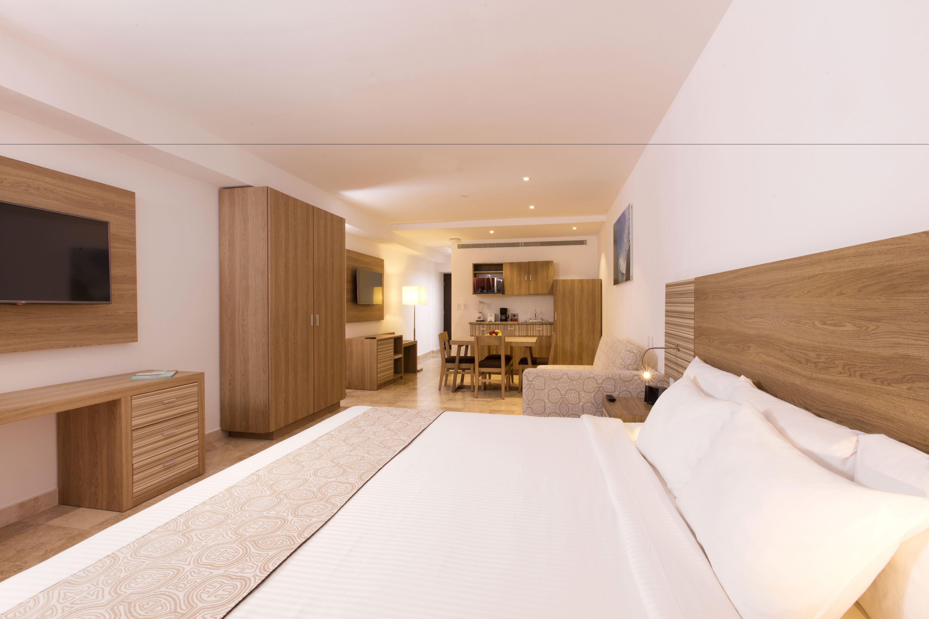 A modern bedroom with a large bed, a TV mounted on the wall, wooden furniture, and a kitchenette in the background.