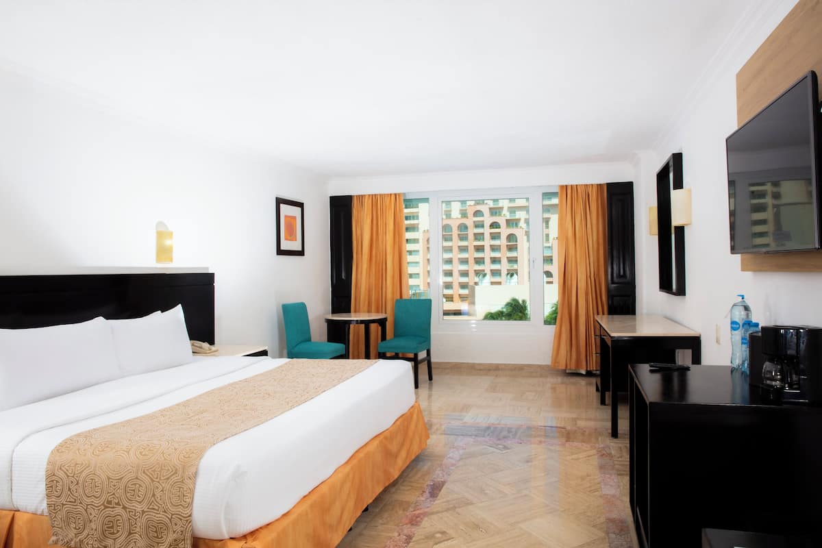 Quarto de hotel com uma cama, uma escrivaninha, duas cadeiras, uma televisão e uma janela com cortinas.