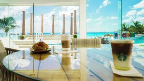 A glass table with a croissant and a Starbucks drink, overlooking a beach with palm trees and a pool. - Krystal Cancun All Inclusive (Cancun)