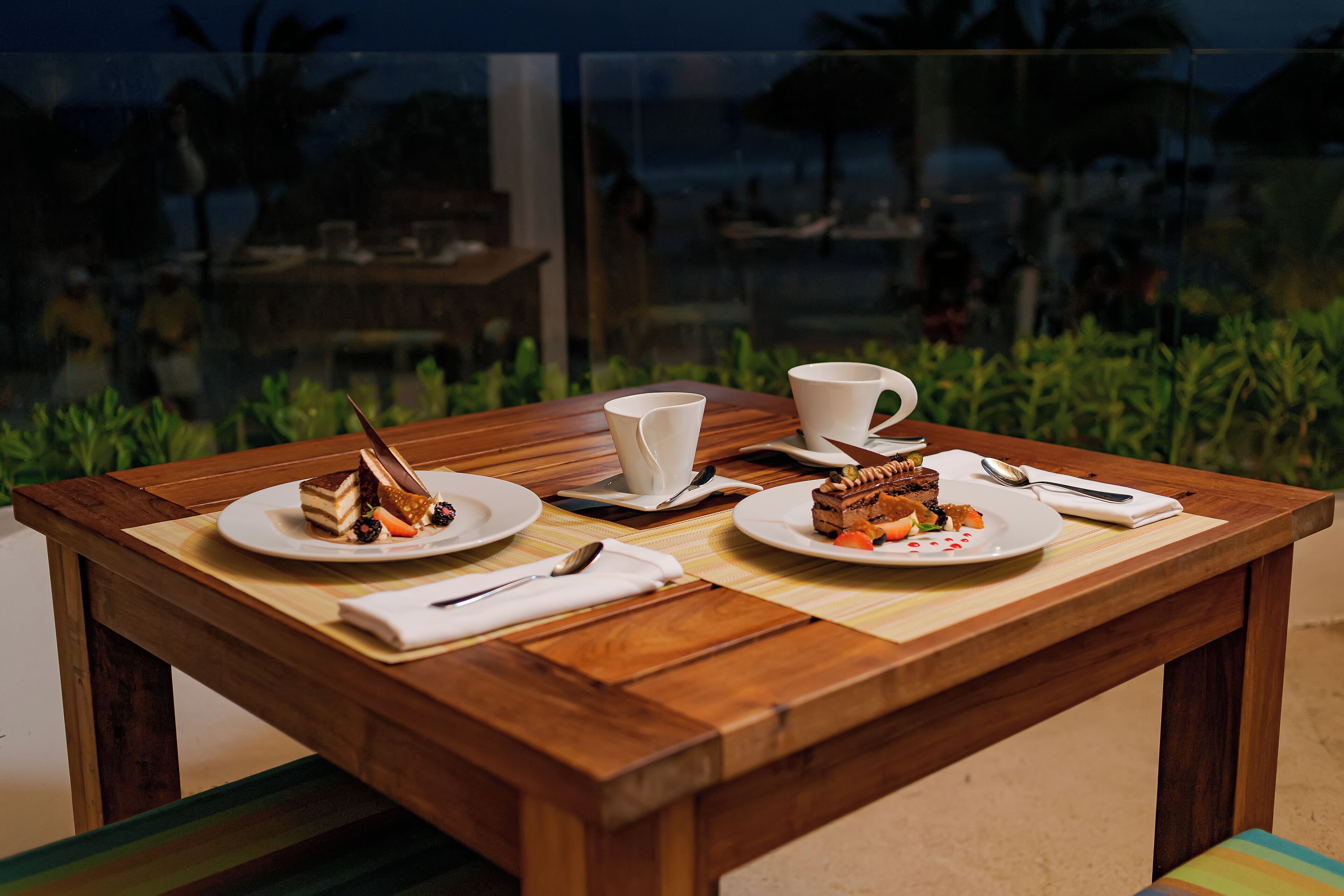 A wooden table with two plates of dessert, a cup, and a spoon, set against a glass window with a view of a beach and palm trees.