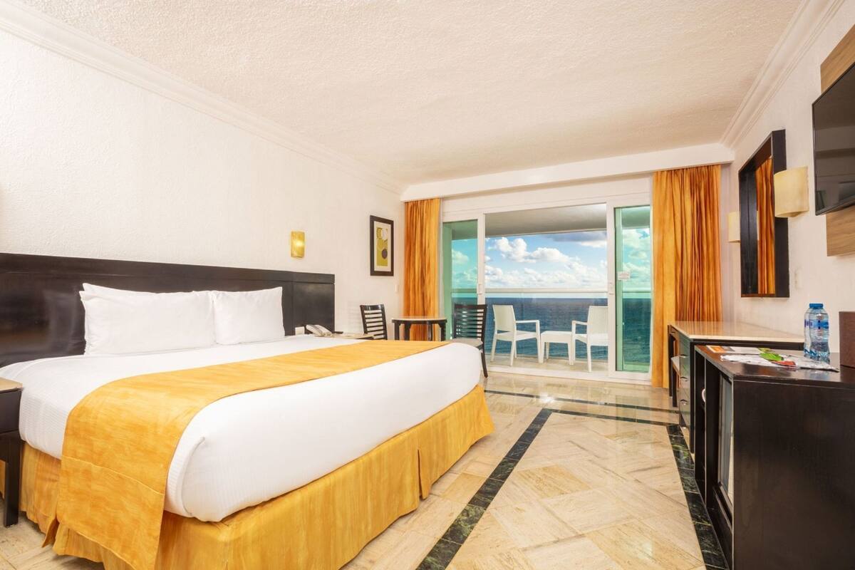 Quarto de hotel com cama, escrivaninha, cadeira, telefone, garrafa de água e vista para o oceano.