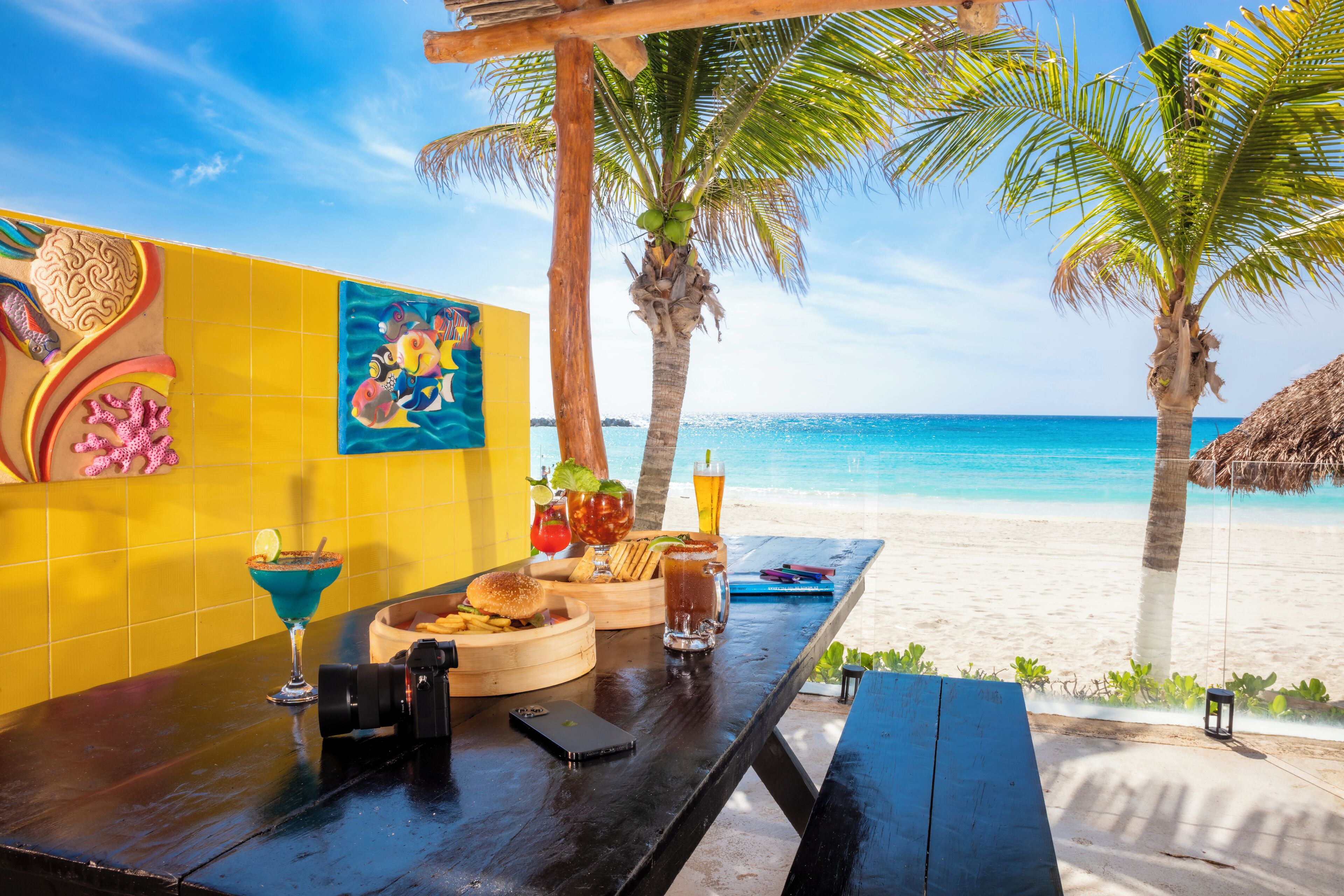 A beachside table with a burger, a drink, and a cocktail, set against a backdrop of a clear blue sea and palm trees.