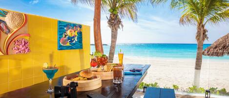 A beachside table with a burger, a drink, and a cocktail, set against a backdrop of a clear blue sea and palm trees.