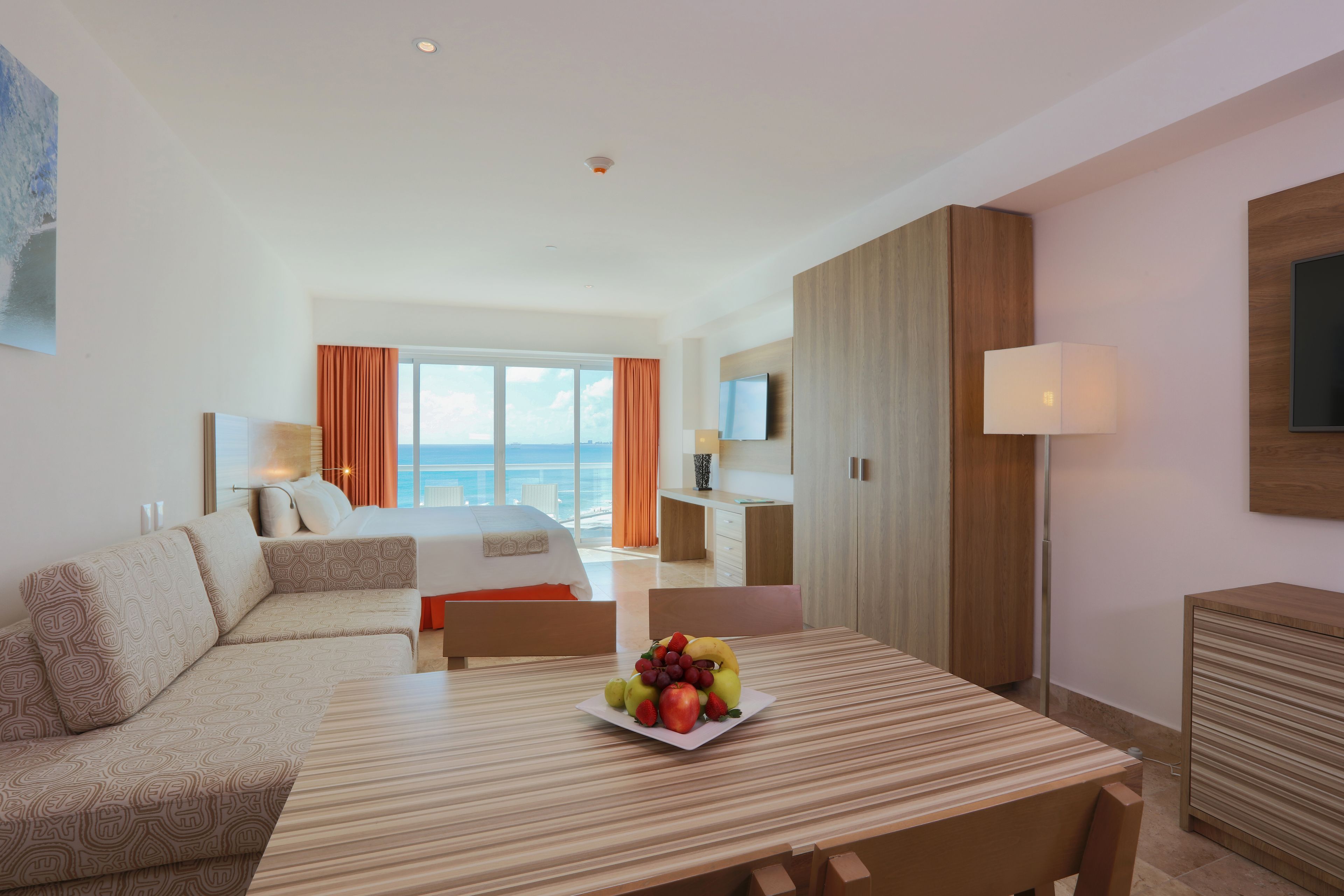 Habitación de hotel con cama, una mesa de comedor con fruta, televisión y vista al océano.