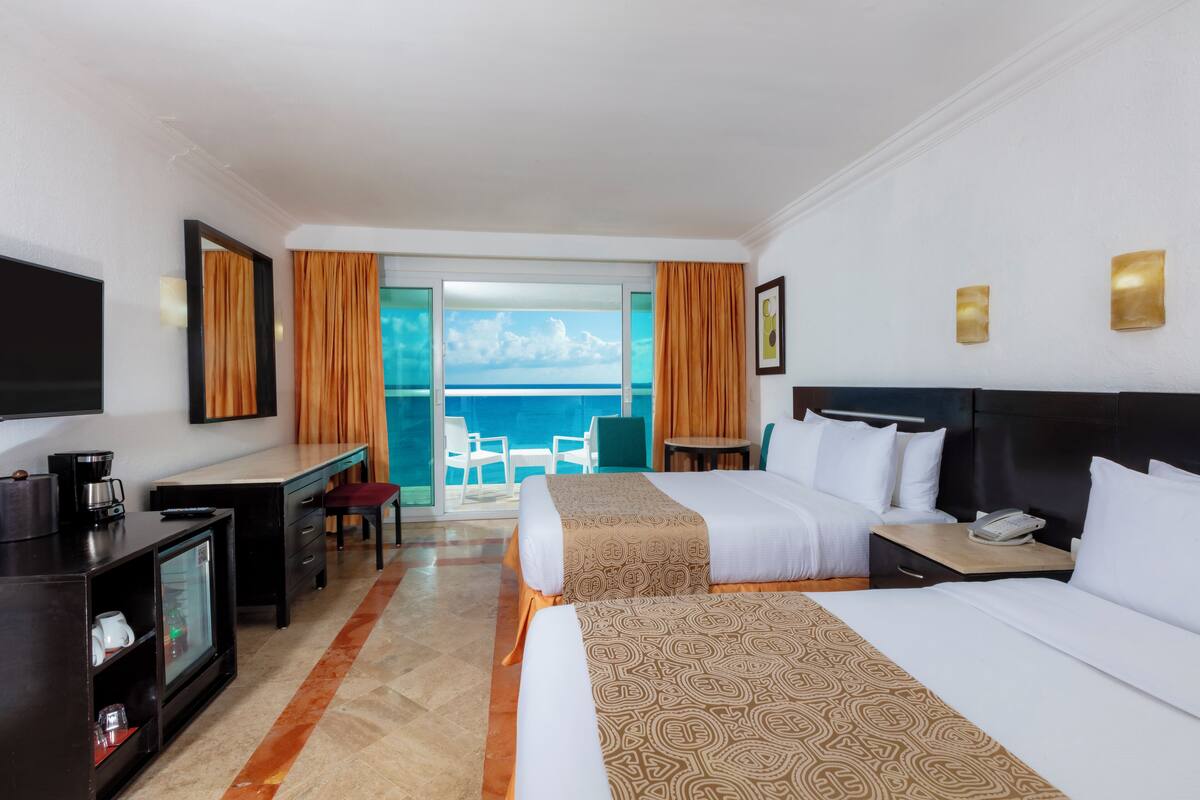 Quarto de hotel com duas camas, uma escrivaninha, televisão e varanda com vista para o oceano.