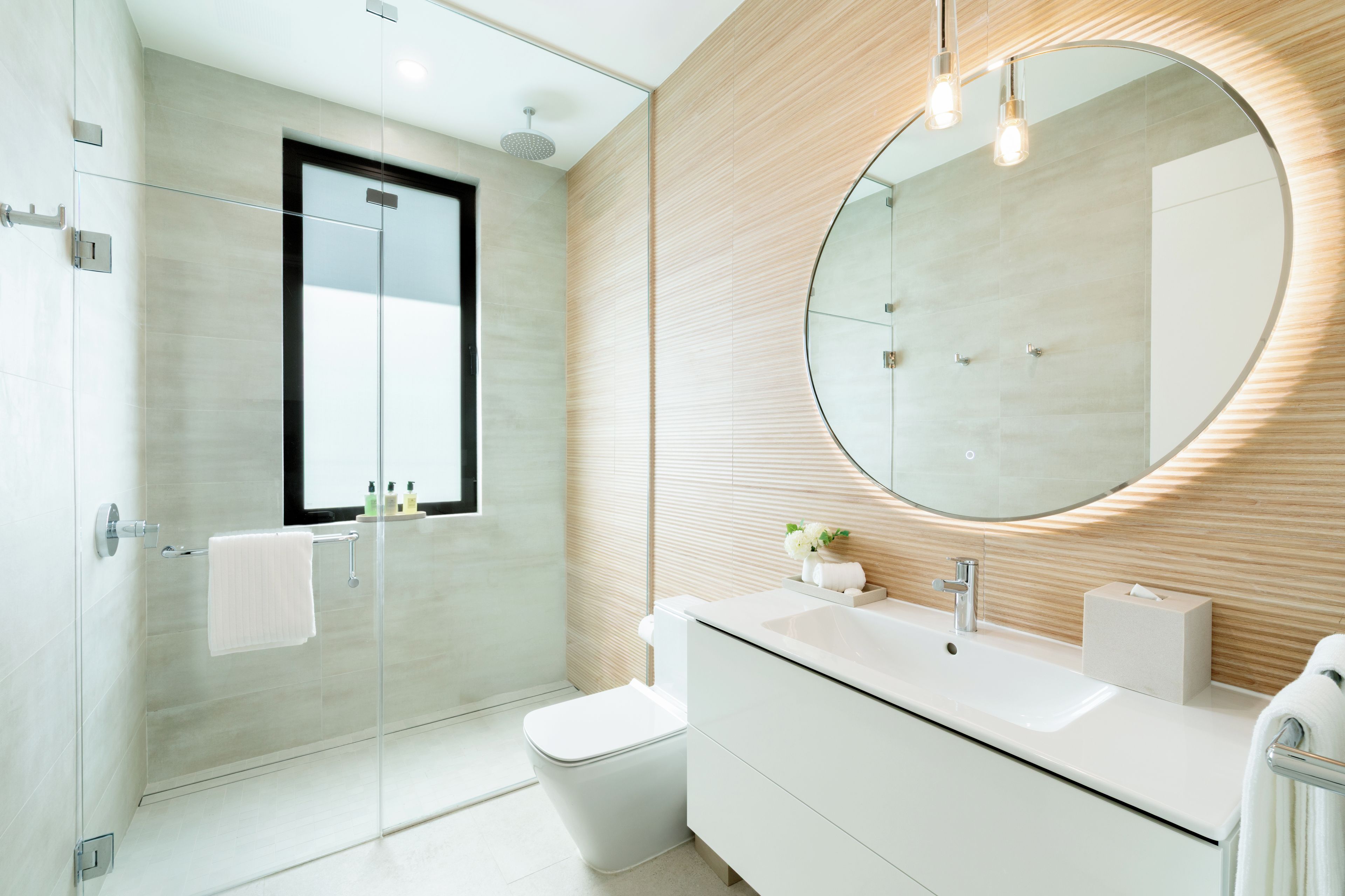 Een moderne badkamer met een douchecabine van glas, een ronde spiegel, een witte wastafel en een toilet.