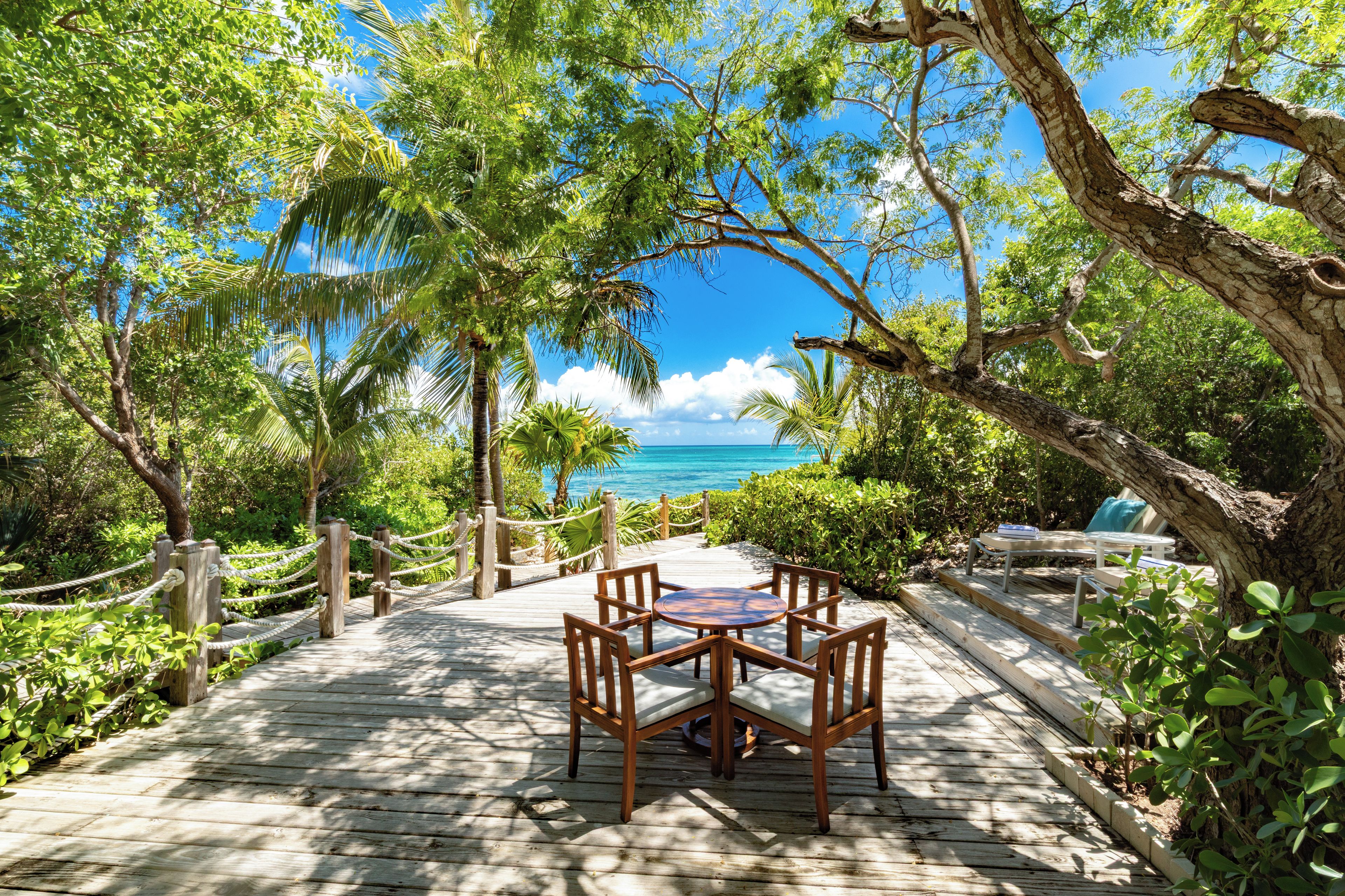 A wooden deck with a table and chairs, surrounded by lush greenery and overlooking a clear blue sea.