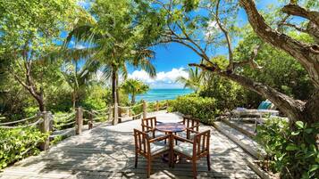 A wooden deck with a table and chairs, surrounded by lush greenery and overlooking a clear blue sea.