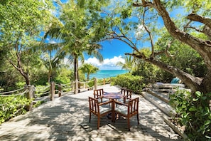 A wooden deck with a table and chairs, surrounded by lush greenery and overlooking a clear blue sea. - Bespoke Villas Turks and Caicos (Providenciales)