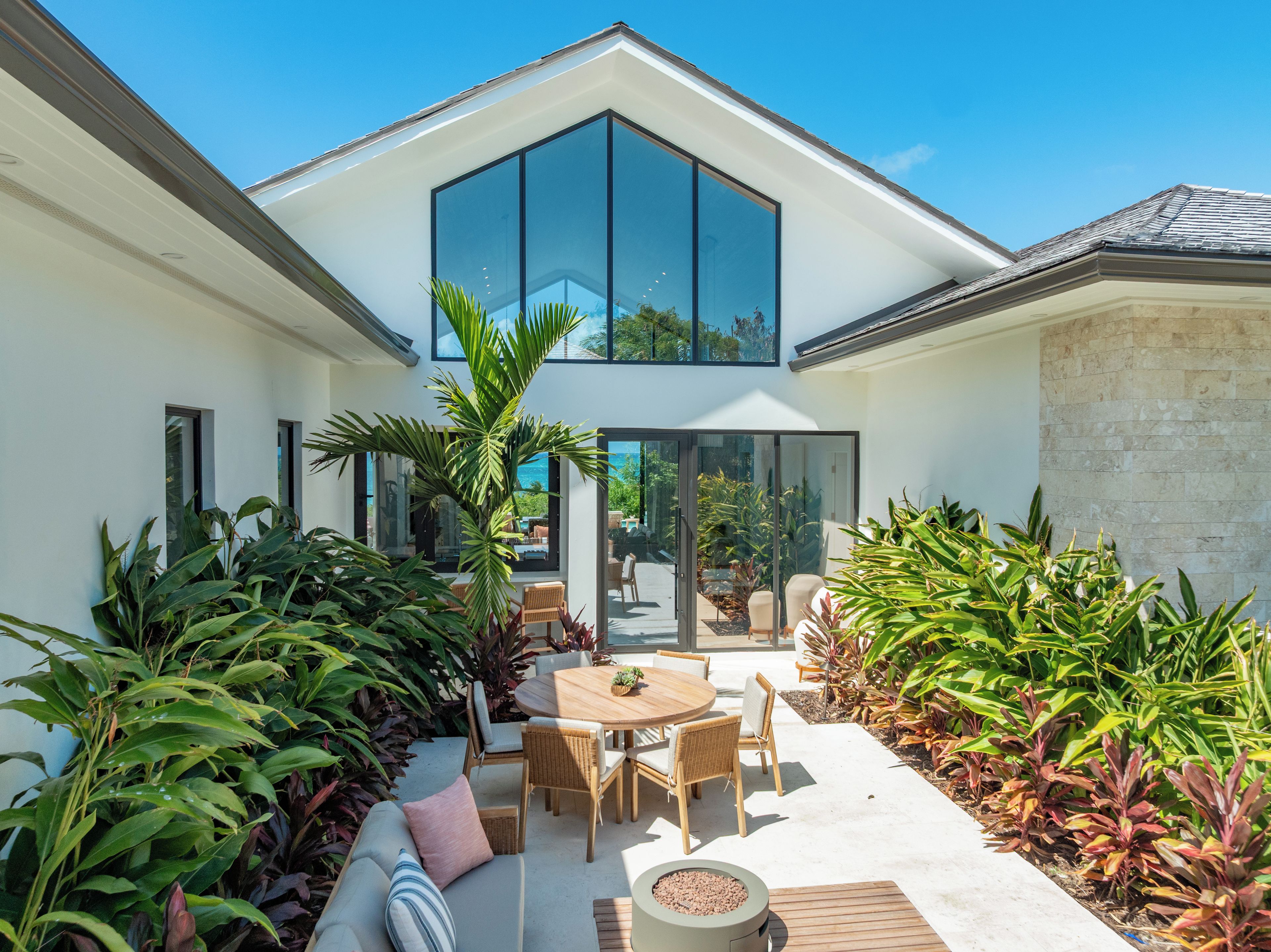 A modern house with a large glass window, a patio with a round table and chairs, and lush greenery.