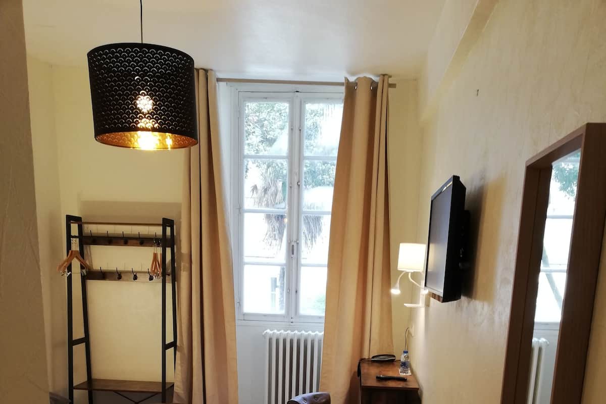 Un dormitorio con una cama, una silla de madera, un escritorio, un televisor, un perchero y una ventana con cortinas.
