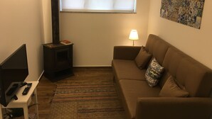A cozy living room with a brown sofa, a small table with a lamp, a TV, a fireplace, and a window with blinds. - Casa do Peso (Peso da Régua)