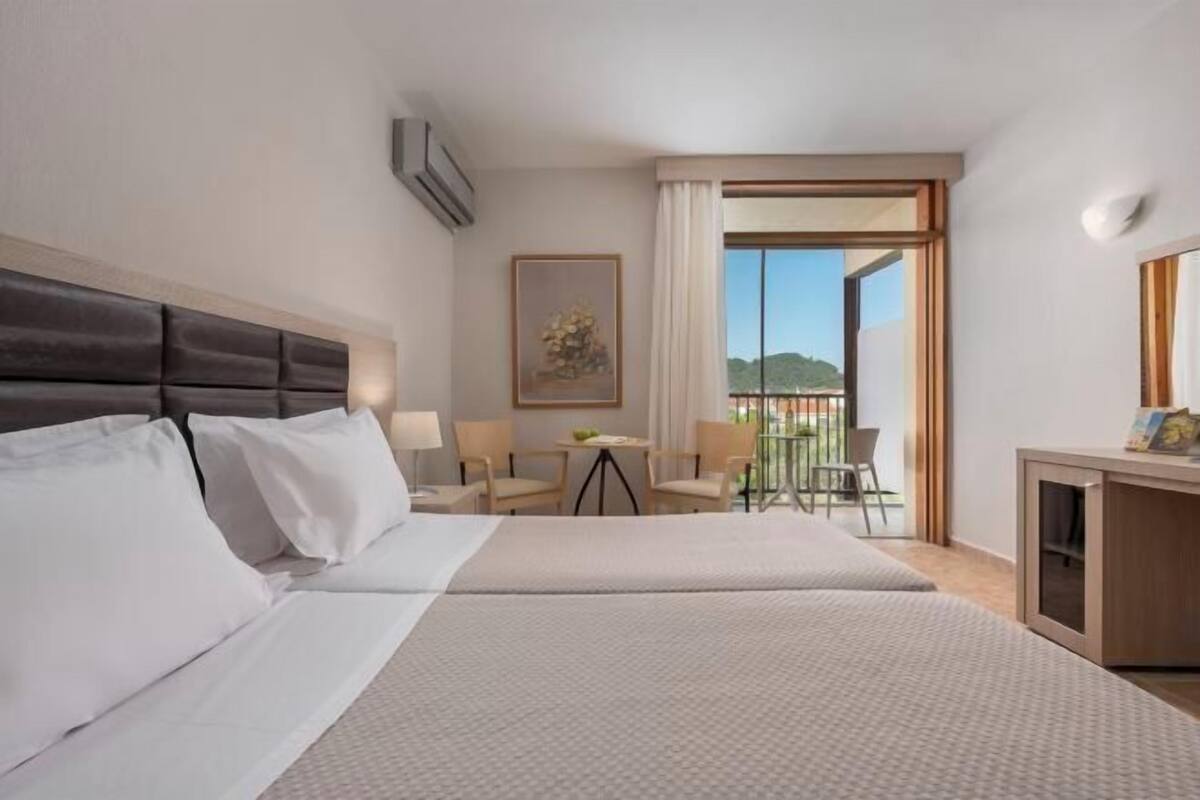 A hotel room with two beds, a desk, a chair, a table, and a balcony with a view.