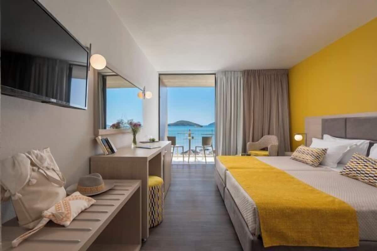A hotel room with a bed, a desk, a chair, a sofa, a TV, and a view of the sea.