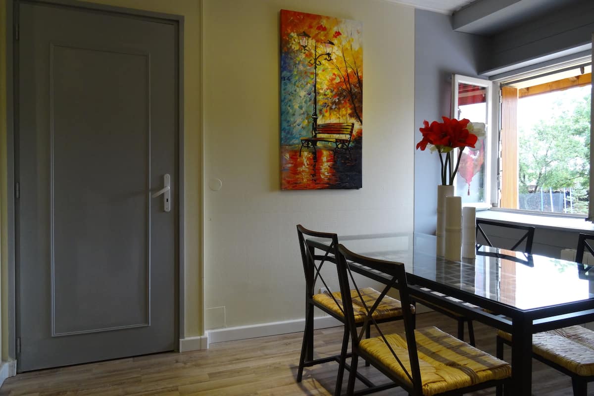 A dining area with a glass-top table, wooden chairs, a colorful painting on the wall, and a window with a view of greenery.