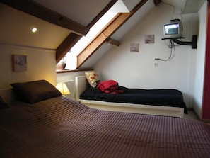 A bedroom with a sloped ceiling, a bed with a striped bedspread, a small bedside table with a lamp, and a television mounted on the wall.